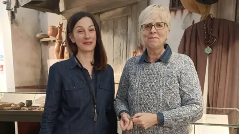 Rugby Borough Council A young woman in a blue shirt stood in front of a medieval display at a museum , next to an older woman with short blonde hair, black glasses and wearing a grey jumper