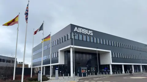 Janine Machin/BBC This is an external picture of a large grey building with two lines of windows with a large "AIRBUS" sign at the top. To the left of the picture four large national flags can be seen. In front of the building is a line of bollards and one person can be seen walking. Behind the building is a grey overcast sky.