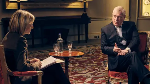 Two people seated opposite each other in upholstered armchairs during a filmed interview in a formal room. A small round table between the chairs holds a glass bottle of water and two drinking glasses. Studio lighting, light stands, microphones and cables are visible around the set. The room has a patterned red-and-cream carpet, decorative wall displays, table lamps, columns and a large framed painting in the background.