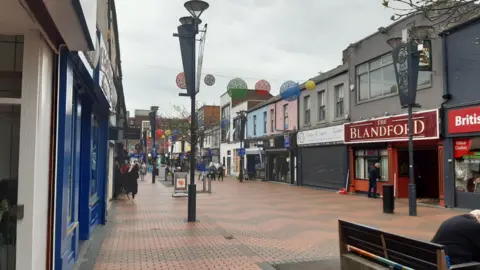 BBC The pedestrianised street is bordered by various shop fronts, including a red shop with a sign that reads: "The Blandford". Red and grey bricks on the road are arranged in a zigzag pattern. Above, large, colourful circular bunting is hung. There are people coming in and out of the shops.