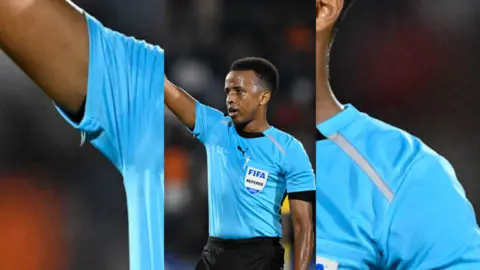 Omar Artan is wearing a light blue referee kit, and has his right arm in the air 