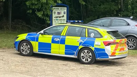 Qays Najm/BBC A police car is in the middle of the picture on some brown concrete. It has chequered blue and yellow pattern, and the front of the car is facing towards the left side of the picture. In the background there are lots of trees, and a sign which says Spanhoe Airfield on it. 