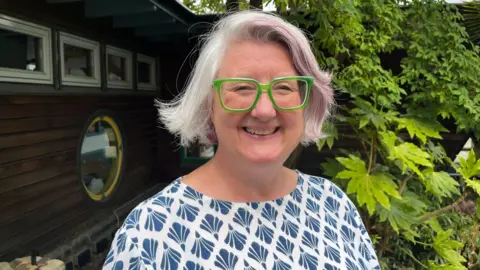 Ollie Conopo/BBC Sarah Beach, outside, smiling and looking at the camera. She is standing by a wooden structure, with trees and bushes to the right. She has short grey hair, is wearing green glasses, and a blue and white top. 