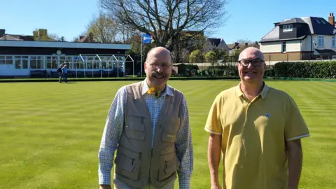 Gregor Macgregor Councillors from Elmbridge Borough Council stand in front of the Walton-on-Thames bowling green. They smile and pose for a photo underneath a clear blue sky bright green lawn, with the bowling club building visible in the background.