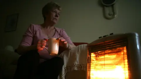 Getty Images A woman sits by an electric heater in a living room. She is wearing a pink jumper and holding a mug.