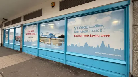 The side of a shop which has large windows entirely covered with blue vinyl which features logos and information about the Stoke Air Ambulance charity. One of the windows features a photograph of a blue helicopter.