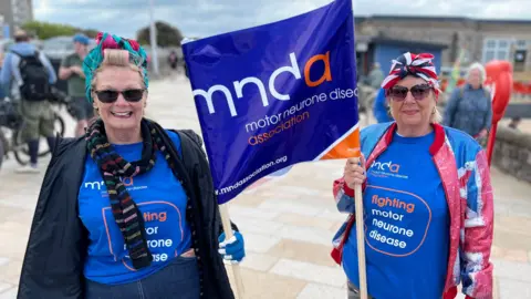 Two women at the tank push event in Weston-super-Mare. They are both carrying a flag and wearing t-shirts with the Motor Neurone Disease Association logo on it.
