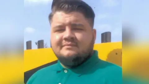 Euan Johnston, a young man with slicked back dark hair and a turquoise green polo shirt, looks into the camera. His eyes are squinting as the sun shines brightly and has a very slight smile. He is standing against a bright yellow wall.