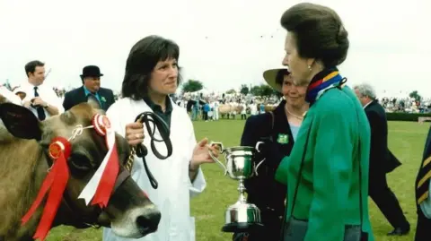 Princess Anne is on the right wearing a green suit and her hair is swept back and pinned. She is facing a woman with dark brown hair and wearing a white lab coat. She is holding a leash which is attached to a cow that has two red rosettes attached to its head.