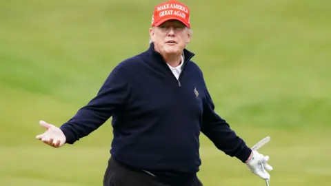 PA A photo of Trump at Turnberry in 2023. He is standing with his arms outstretched, holding a golf putter. He is wearing one white glove. He has a blue fleecy sweater on and a red MAGA hat. The background is the green of the golf course. 