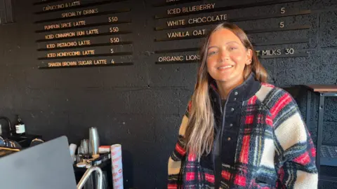 BBC Riot Cycles owner Claire Reynolds standing in front of a black wall with yellow lettering showcasing the prices of her items. She is wearing a woolly cream, red, and black coat. She is smiling looking at the camera. In the background there are plastic cups, coffee jars, and a roll of adhesive window protector. 