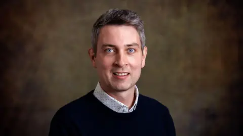 Dr Rory Jones A man in a blue jumper with a shirt underneath.