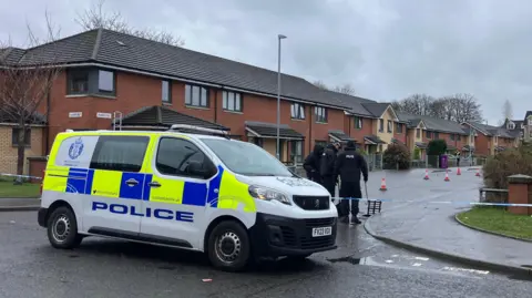 BBC A police van block the entrance to a residential street which has been sealed off with blue and white police crime scene tape