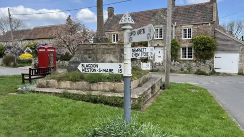 An old-fashioned white fingerpost on a village green in a chocolate box-type village with a red telephone box and bench next to a war memorial and a couple of stone houses in the background. Places on the fingerpost include Weston-super-Mare and Blagdon