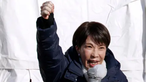 Sanae Takaichi speaks into a microphone with her fist raised. She's wearing a dark blue puffer coat