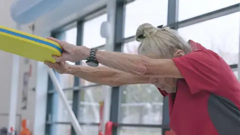 EVERYONE ACTIVE LEISURE Jennifer Brackley, with grey hair tied up in a ponytail, wears a red sports shirt. She has her head down and her arms outstretched in front of her. She is holding a yellow and blue pool float in her hands. She is by the window of a swimming pool.