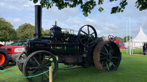 Luke Deal/BBC A vintage ploughing vehicle parked in a grass field. It is black and has large wheels as well as a large vertical pipe from its engine on the front.