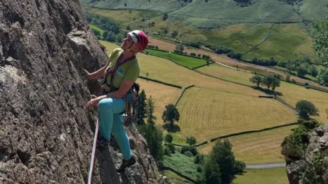 Stella Adams Stella Adams, climbing in the Lake District. She is climbing a ribbed rock face which is very exposed onto the patchwork of fields and hedges in the valley below her. She has one foot perched higher up and the other leg straight, and does not appear to be holding on with her hands. The rock has a purple-tint and the valley below her is very green. She is wearing a pink climbing helmet, a green t-shirt and turquoise blue trousers. 