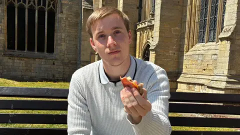 James Quirke, a man with blonde hair, wearing a light-coloured jumper and jeans, sits on a bench with part of a church visible in the background. He is holding out a half-eaten bacon sandwich, showing it to the camera. 