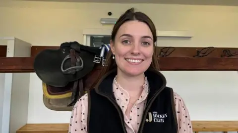 A woman smiles as she stands in the tack room at a racecourse. She has long dark-brown hair and is wearing a black gillet with "Jokey Club" logo over a pink blouse. Behind her, saddles rest on a wooden beam.