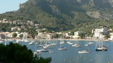 BBC A bay filled with boats with large rocky cliffs covered in greenery in the background in Majorca