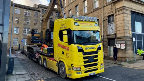 A yellow demolition excavator on the back of a bright yellow truck.