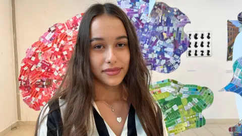 Nicola Haseler/BBC A young woman with long, straight brown hair smiles at the camera. She is wearing a black and white striped blouse and is standing in front of a colourful cardboard sculpture of a brain.