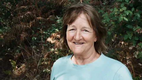 John Fairhall/BBC A woman with brown, shoulder-length hair and a fringe swept to either side of her face, looks directly at the camera. She wears a light blue T-shirt with the chain of a thin silver necklace party visible. The background shows dense green and brown forest foliage, including ferns and leafy branches.