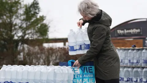 PA Media A woman wearing a dark coat is lifting a multi‑pack of bottled water from a pallet outdoors and placing it into a reusable shopping bag. Rows of similar water packs are stacked beside them, with trees and a building in the background.