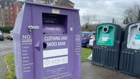 A purple clothing and shoes donation bin ata car park, with bottle bank bins and parked cars nearby. There is a large block of flats in the background.