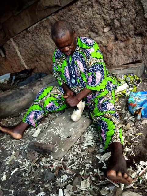Molara Wood Kasali Akangbe Ogun, vestido com uma roupa verde colorida, está sentado no chão e fazendo uma escultura