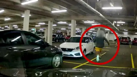 A man in blue sneakers is circled in red, running through a car park.