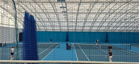 Dumfries Tennis Club Two indoor tennis courts, viewed through netting. There is a doubles game on the right hand court and a singles match on the left. 
