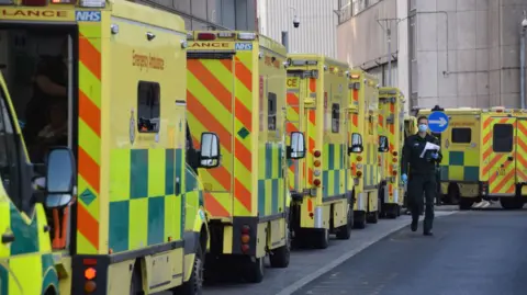 A line of yellow and green ambulances parked on a road. A paramedic wearing a mask is heading towards the camera. There is a right arrow sign in the background, as well as a tall white building.