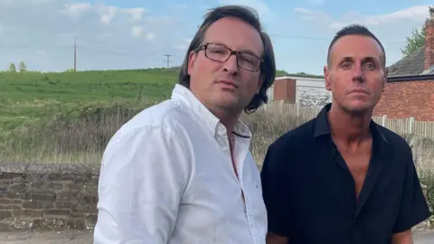 Tom MacDougall/BBC A man with dark hair wearing a white shirt and dark framed glasses stands next to a man with short dark hair wearing a black shirt. Behind them is a low wall and then fields beyond.