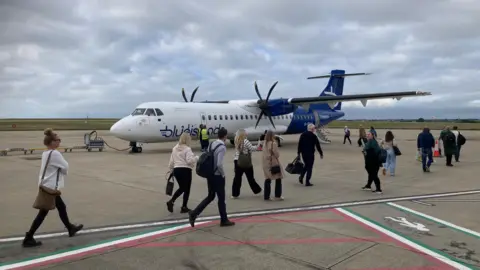 BBC Passengers boarding a Blue Islands flight from Jersey to Birmingham 14 October 2024. The aircraft is mostly blue and white. Passengers are carrying their luggage while walking to the back end of the aircraft where they will board the plane. It is a grey day. 