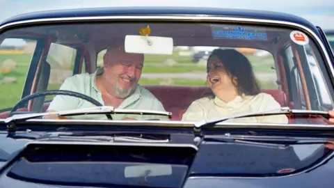 ITV Cymru Wales / BBC Cymru Wales Jones and Speirs in an old navy vintage car laughing with each other. Jones, on the right, is in the passenger seat and is laughing whilst looking at Speirs. She wears a cream jumper and has brown hair. Speirs, is on the left, and wears a light green t-shirt and is looking down while laughing.