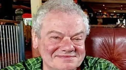 Family Photo Brad smiling and wearing a green plaid shirt sitting in a leather chair. A bar can be seen behind him. He has short silver curly hair