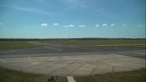 An airport runway strip spans across the whole picture. There is a circle at the end of a runway where planes turn. In the foreground are trees.