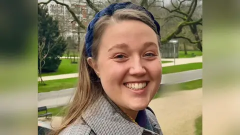 British Medical Association Alice is stood outside with green spaces, benches and buildings in the background. She is wearing a checked jacket and a navy blue head-band. She has long brown hair and is smiling at the camera. It is a head and shoulder shot of her.