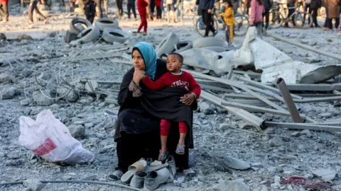 Getty Images Seorang wanita, duduk di reruntuhan di Gaza, dengan seorang anak laki-laki di pangkuannya. Ada juga orang di latar belakang. 