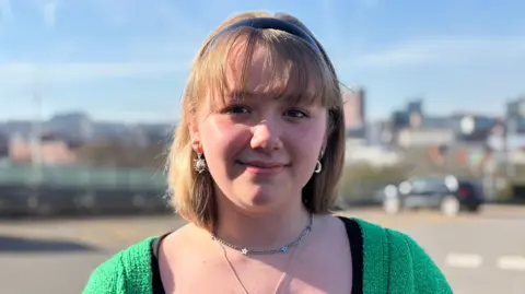 Simon Thake/BBC 16-year-old Emmie is outside Sheffield College in the sunshine and is smiling. She is wearing a black top and a green cardigan.