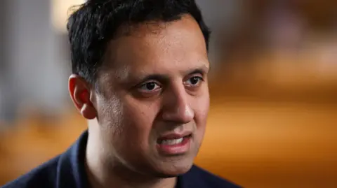 Anas Sarwar has dark hair and is wearing a dark t-shirt
