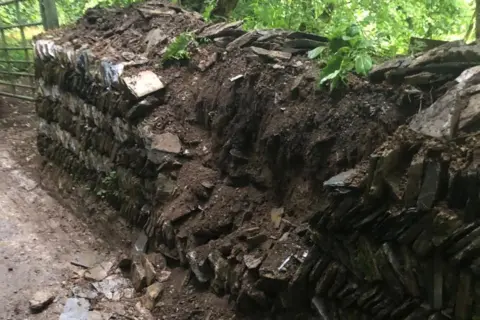 A stone wall lining a road. Large parts of the centre of the wall have been damaged and are falling in to the road.