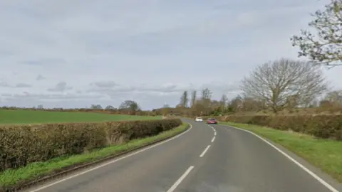 A country road with white lines down the middle and at the edges, with a small verge and low hedges on both sides. 