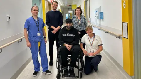 Prof Kirk Levins Five people smile at the camera. On the left, a shorter man with dark hair wearing blue hospital scrubs. On his right, a taller man with dark hair wearing a navy jumper and black jeans.  In the middle, a man wearing a black hoodie and black tracksuit bottoms sits in a wheelchair. On his right, a woman with dark hair stands wearing a black and white patterned top. On her right, a blonder woman in a white t-shirt crouches next to the man in the wheelchair. The five people are in a long white corridor which is lit with harsh lighting. 