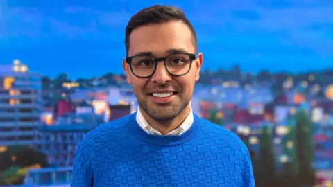 BBC Dan Miller looking and smiling at the camera with a blurred skyline of a city. He's wearing glasses and a blue jumper over a white shirt.