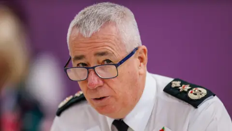 PA Media Jon Boucher is wearing the white shift with black shoulder patches of the PSNI. He wears a black tie, his glasses are blue and hair is grey, short and matted. 