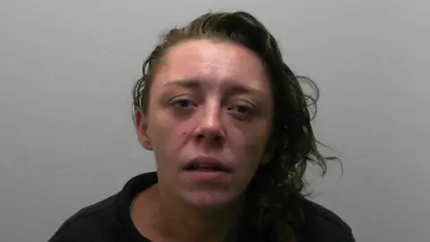 A custody photo of a woman sitting against a plain light‑coloured wall, wearing a black zip‑up jacket.