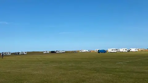 More than 10 white caravans have parked up along the edges of a football pitch. Two vans, one grey and one navy, are parked with the caravans. There are tyre marks in the grass heading towards where the camp has been set up.